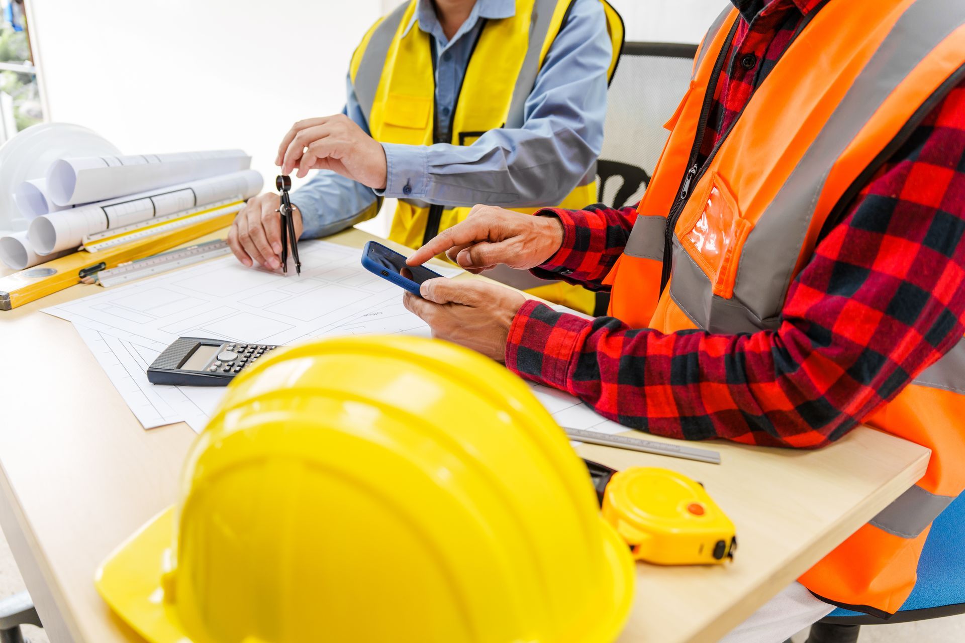 Two construction workers reviewing blueprints, using a calculator and compass, wearing safety vests and hard hats.