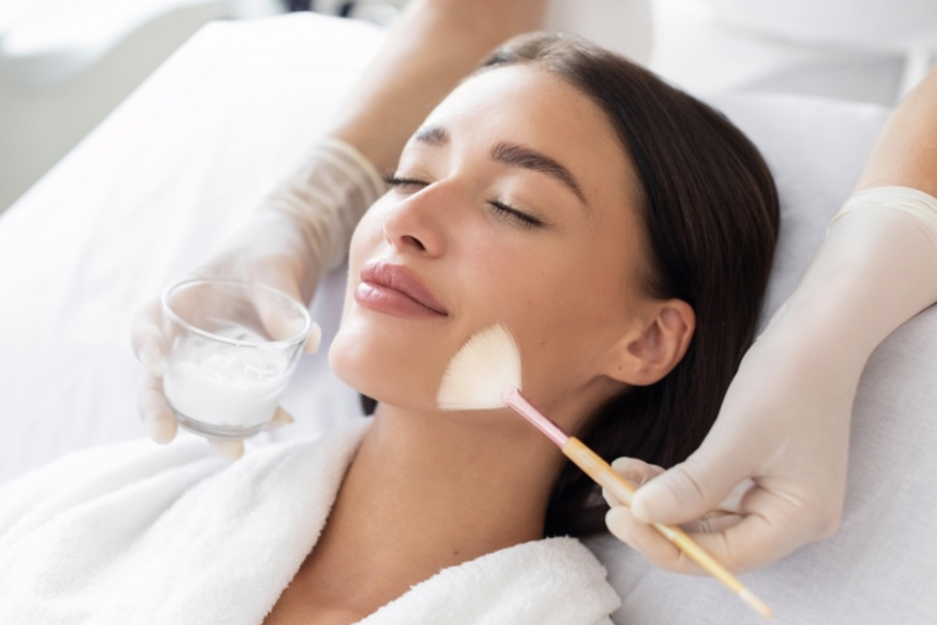 Woman receiving a facial chemical peel at a spa; an aesthetician applies a substance to her cheek with a brush.