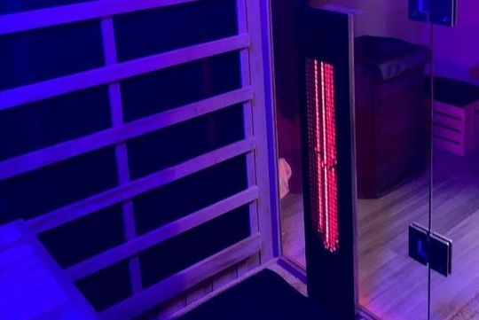 A dark, purple-lit sauna interior. A red heating panel is visible near the glass door. infrared sauna. mid, near and far infrared.
