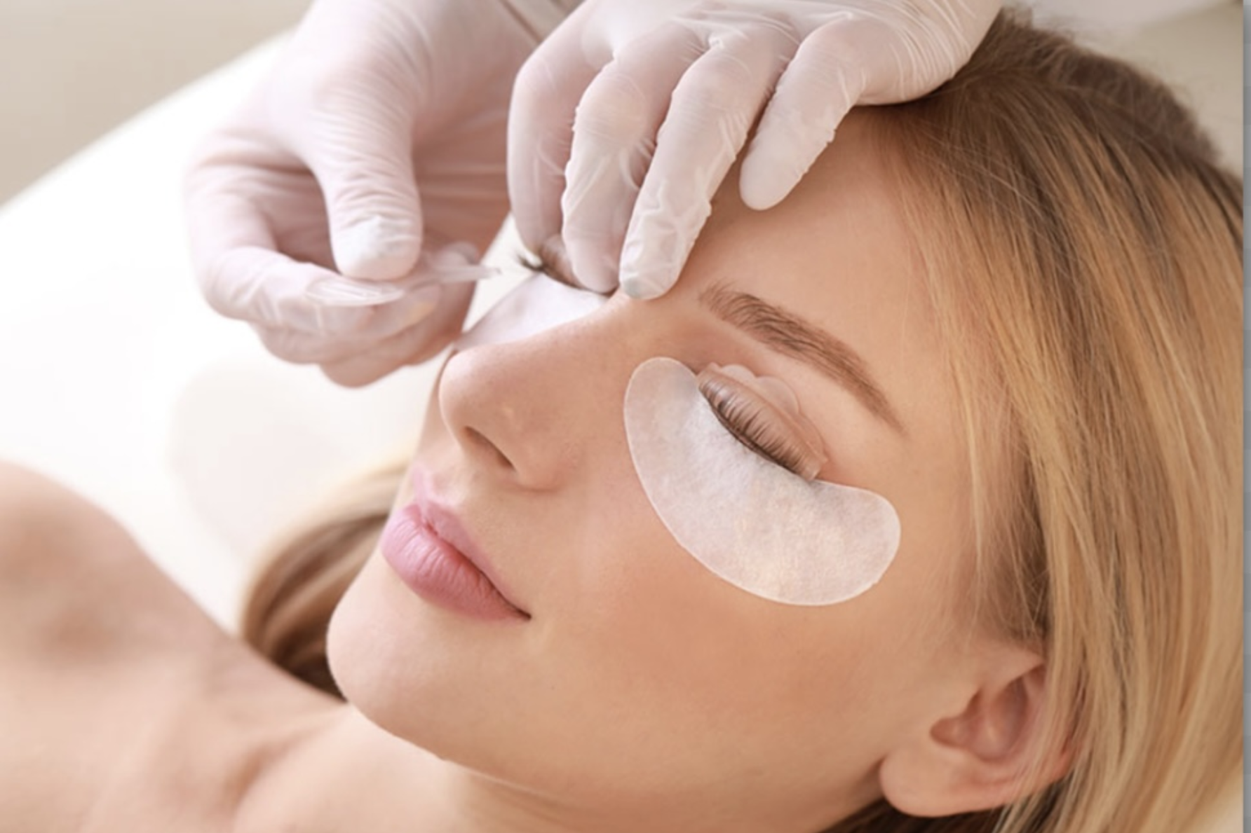Woman receiving a lash lift; technician in gloves applies pads, then product to lashes in a salon setting.