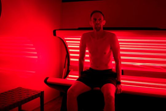 Man sits shirtless in a red light therapy booth; the room is dimly lit. red light bed. 2 red light frequencies and 3 infrared frequencies.