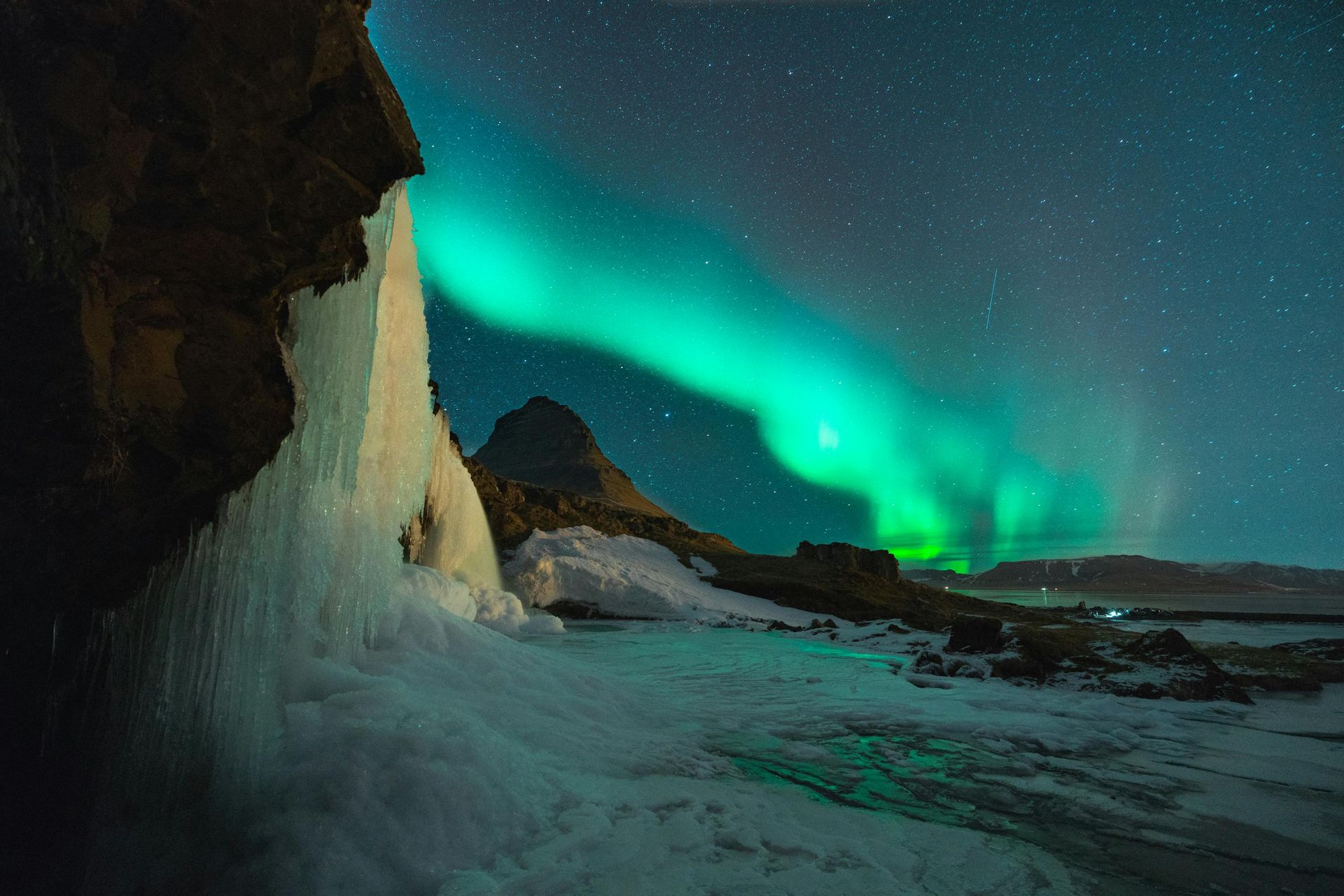 Aurora borealis illuminates the night sky over a frozen waterfall and mountain in Iceland.