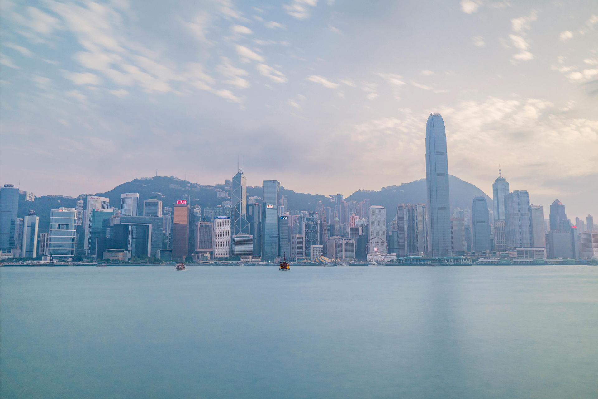 Hong Kong skyline with many skyscrapers, seen across a calm, blue-green sea under a cloudy sky.
