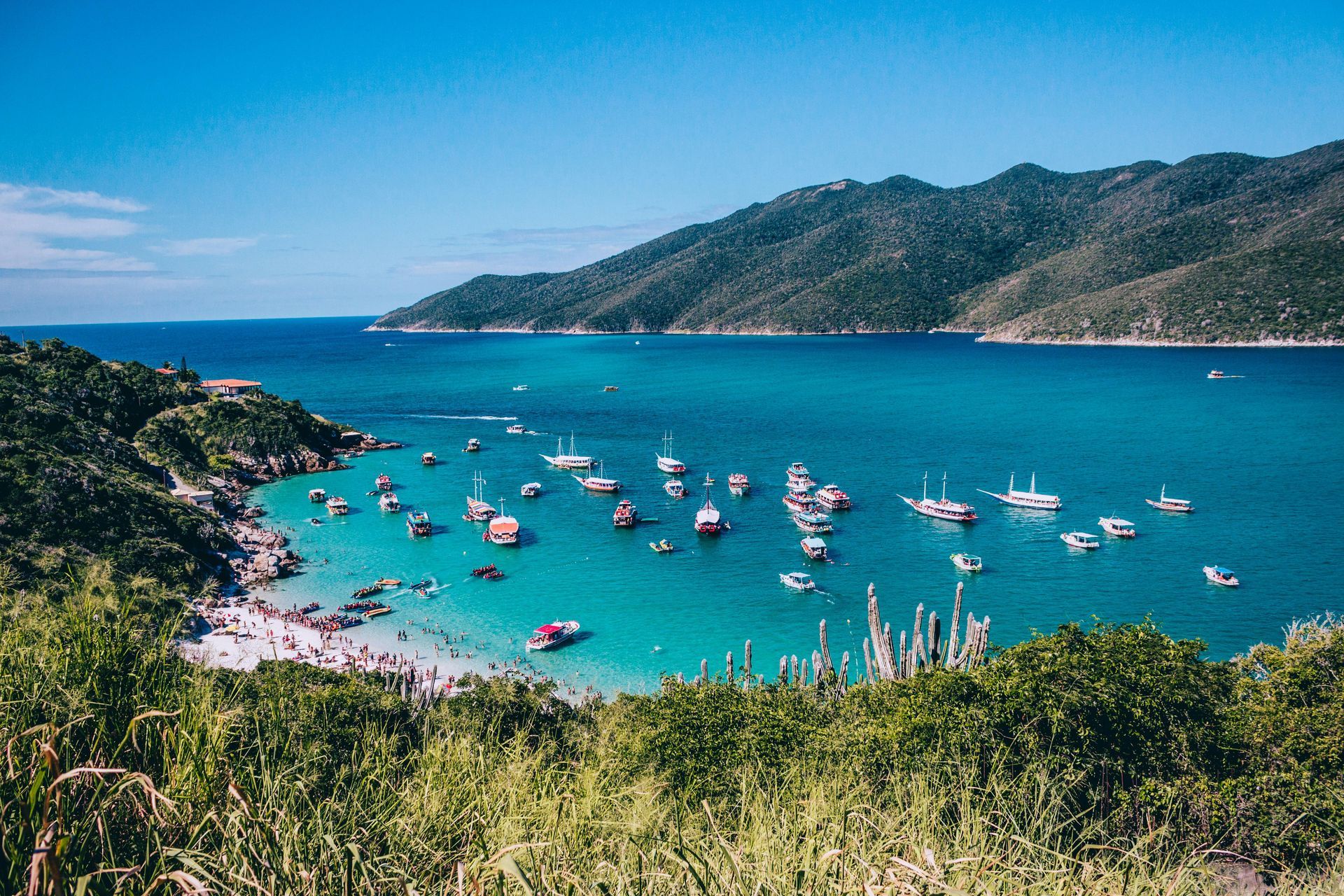 Boats in turquoise water, beach, and lush green hills under a bright blue sky.