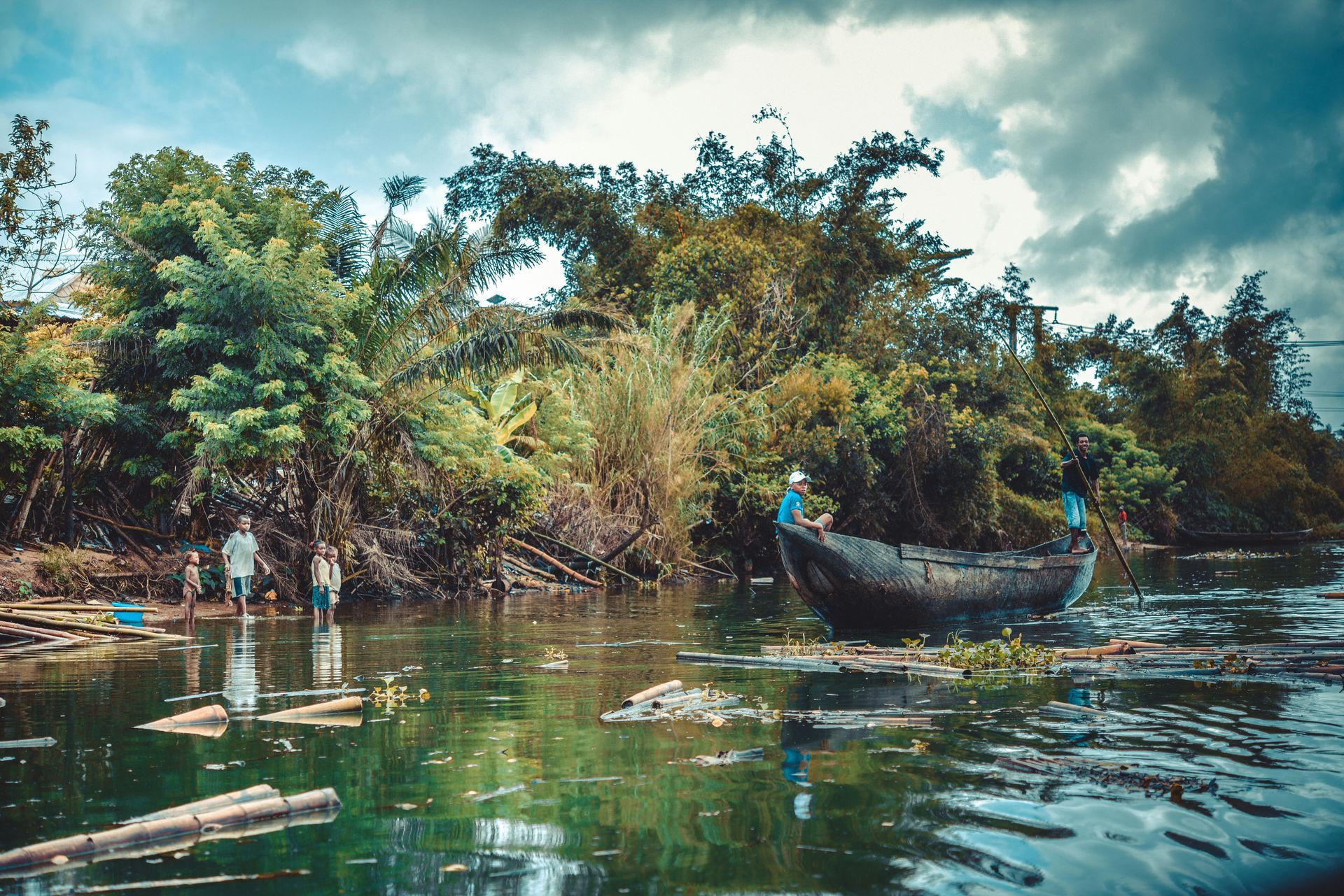 A dark wooden boat sits in murky water, with trees and people on a muddy bank under a cloudy sky.