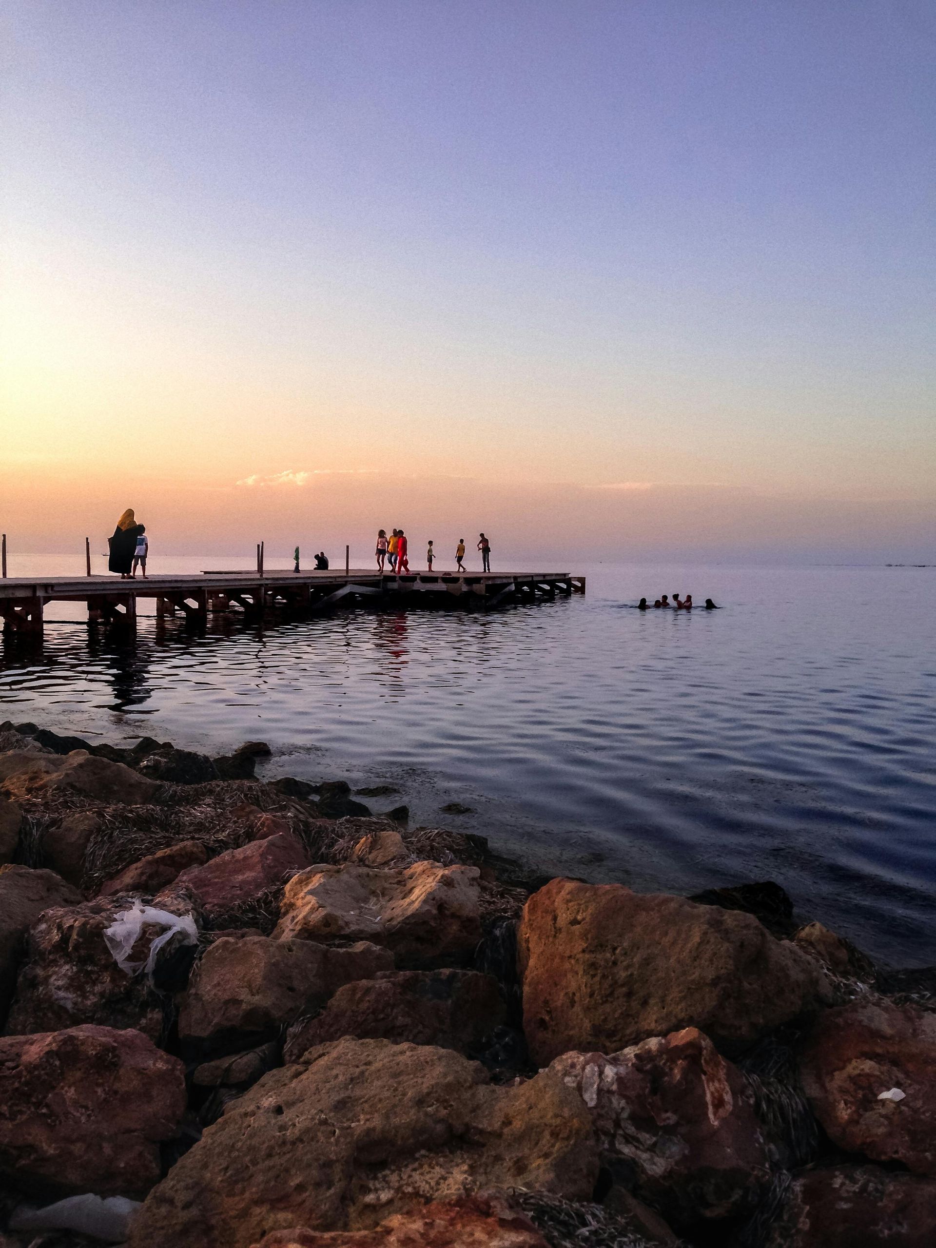 Sunset over calm water; people on a pier and swimming. Rocks in foreground.