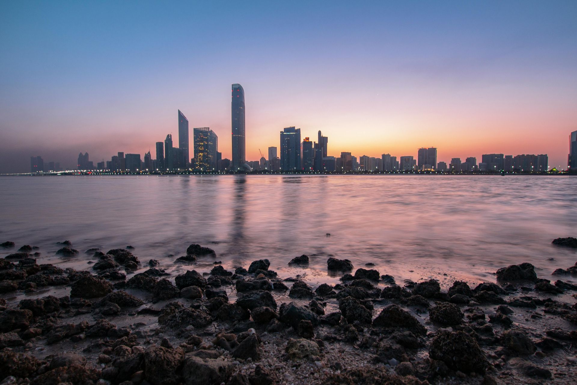 Skyline at dusk over water, rocks in foreground, buildings silhouetted against pink and blue sky.