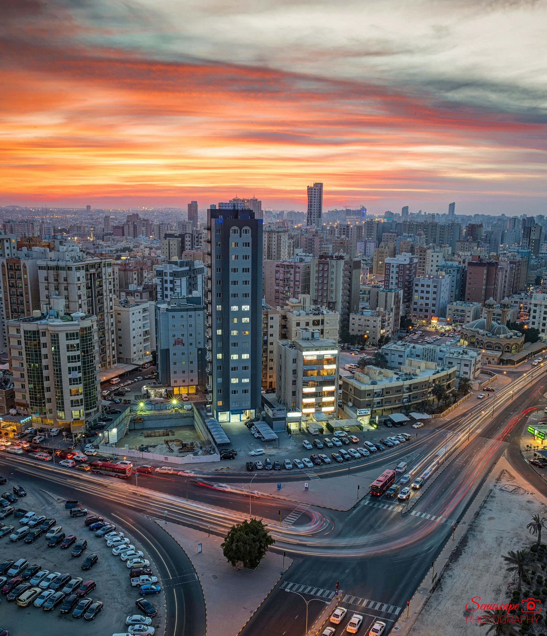 Cityscape at sunset with colorful sky, buildings, roads, and parked cars.