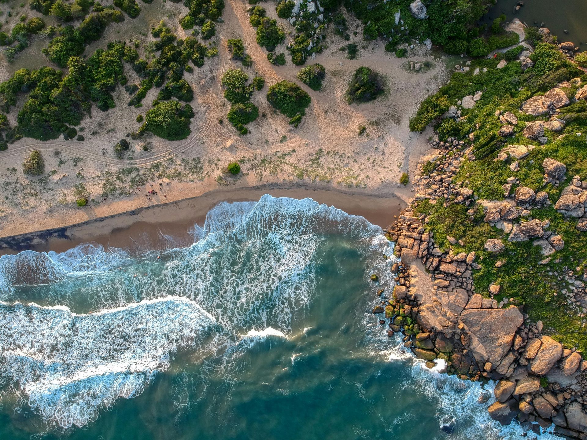 Aerial view: Ocean waves crashing on a sandy beach, bordered by rocks and lush green vegetation.