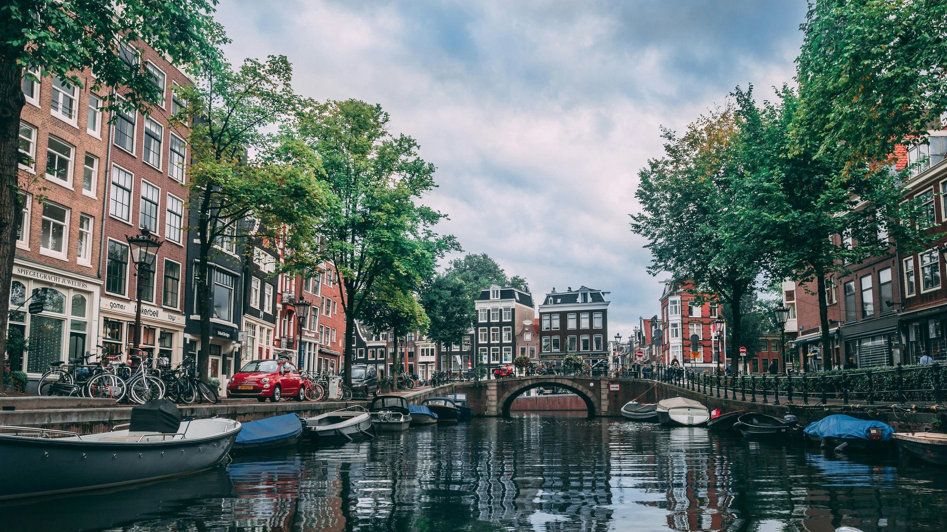 Canal in Amsterdam lined with buildings, boats, and a bridge under a cloudy sky.