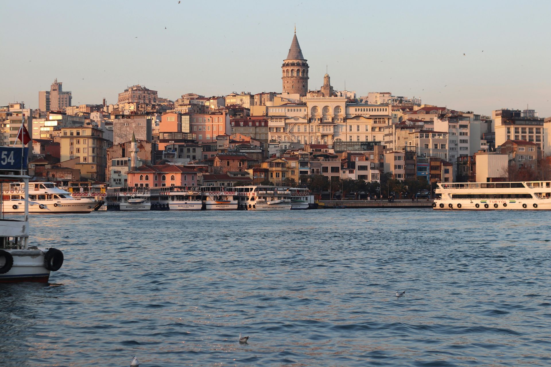Galata Tower overlooks buildings by water with boats at sunset.