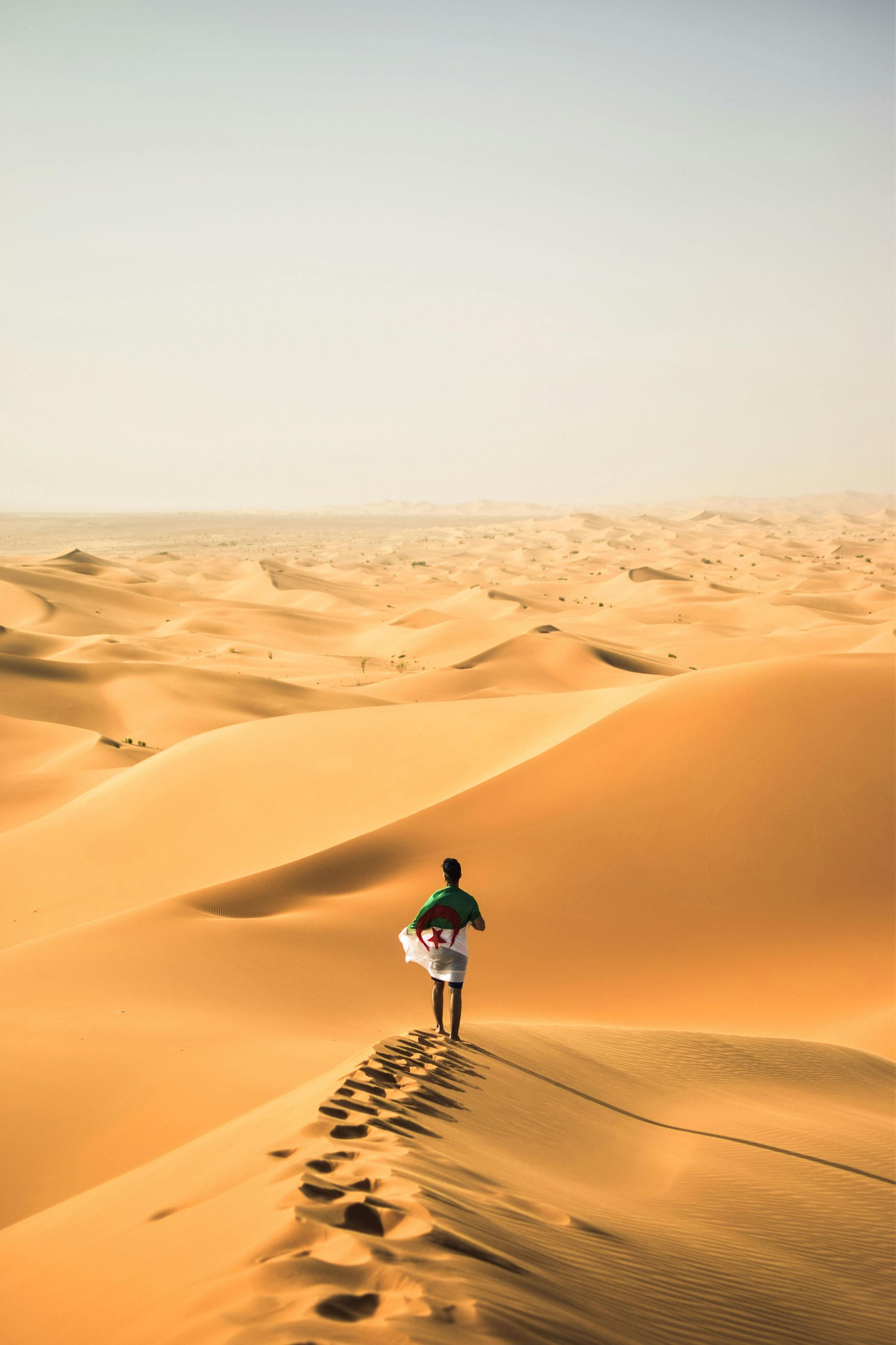 Person walking on a sand dune in the desert, leaving footprints, holding a white cloth, sunny day.