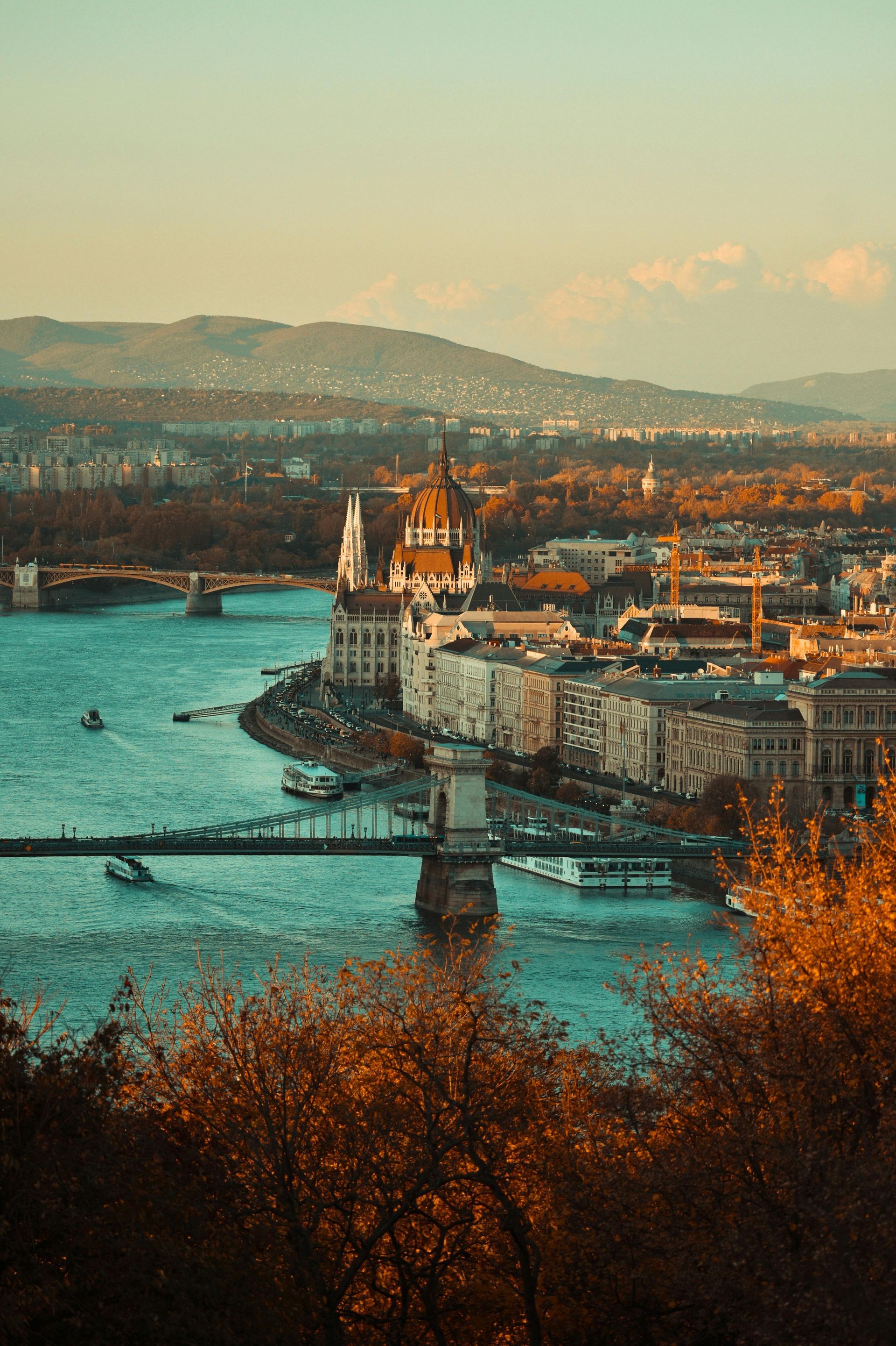 Budapest cityscape: Danube River, Parliament building, Chain Bridge, autumn colors, mountains in the background.