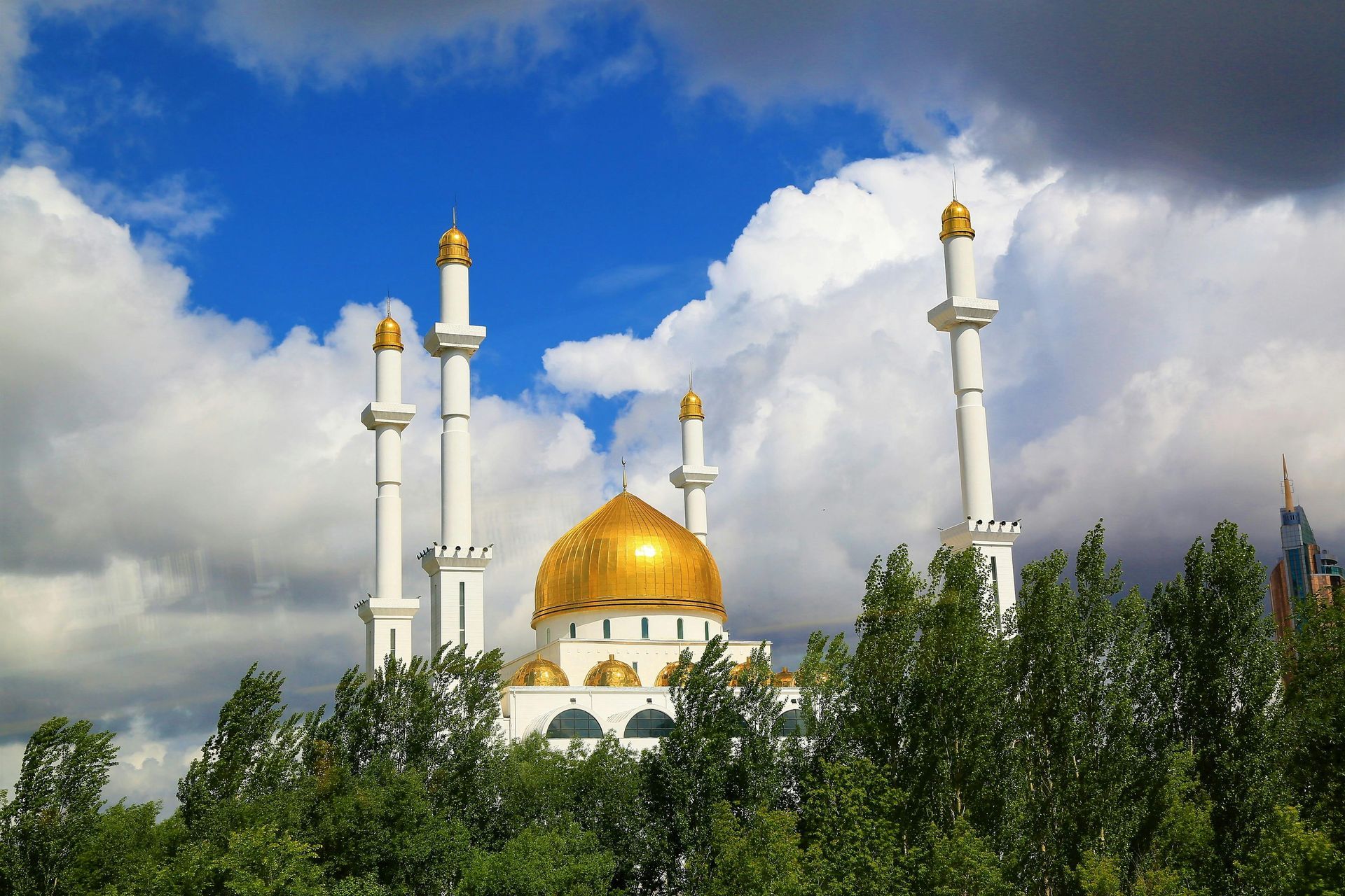 Golden-domed mosque with white towers, against a blue sky with puffy white clouds, seen from behind trees.
