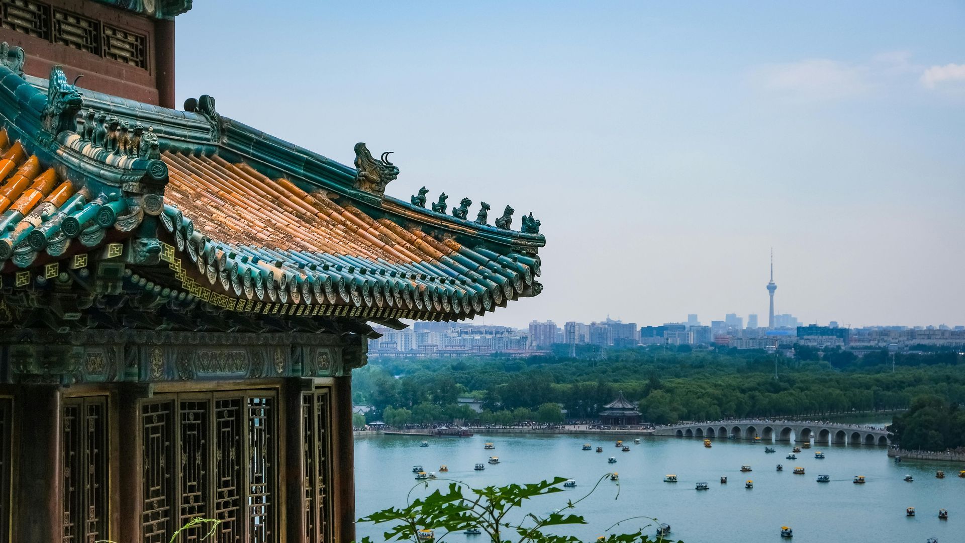 Traditional Chinese building overlooking a lake and city skyline. Teal roof, boats, and distant trees.