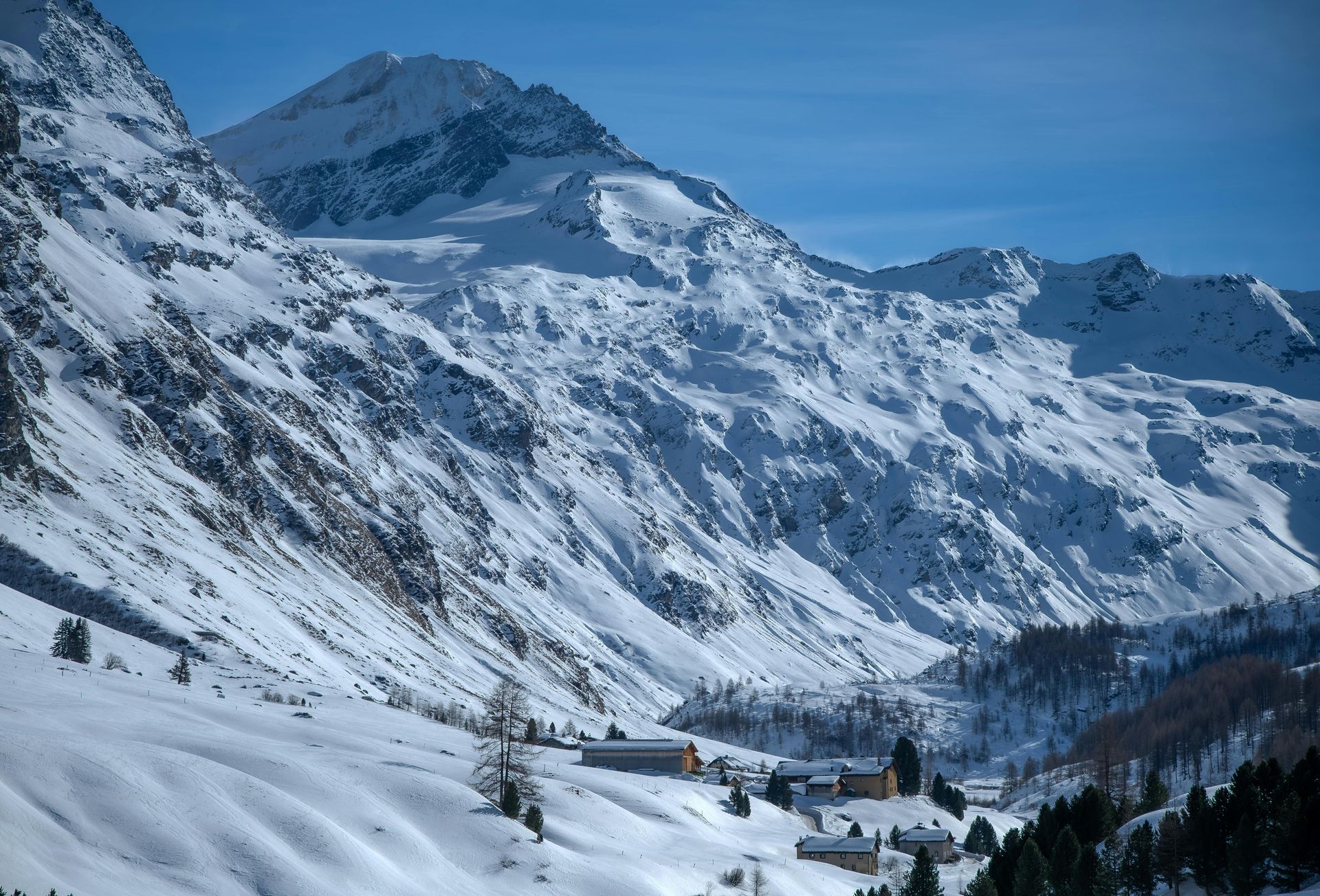 Snow-covered mountains, a valley with buildings, and a clear blue sky.