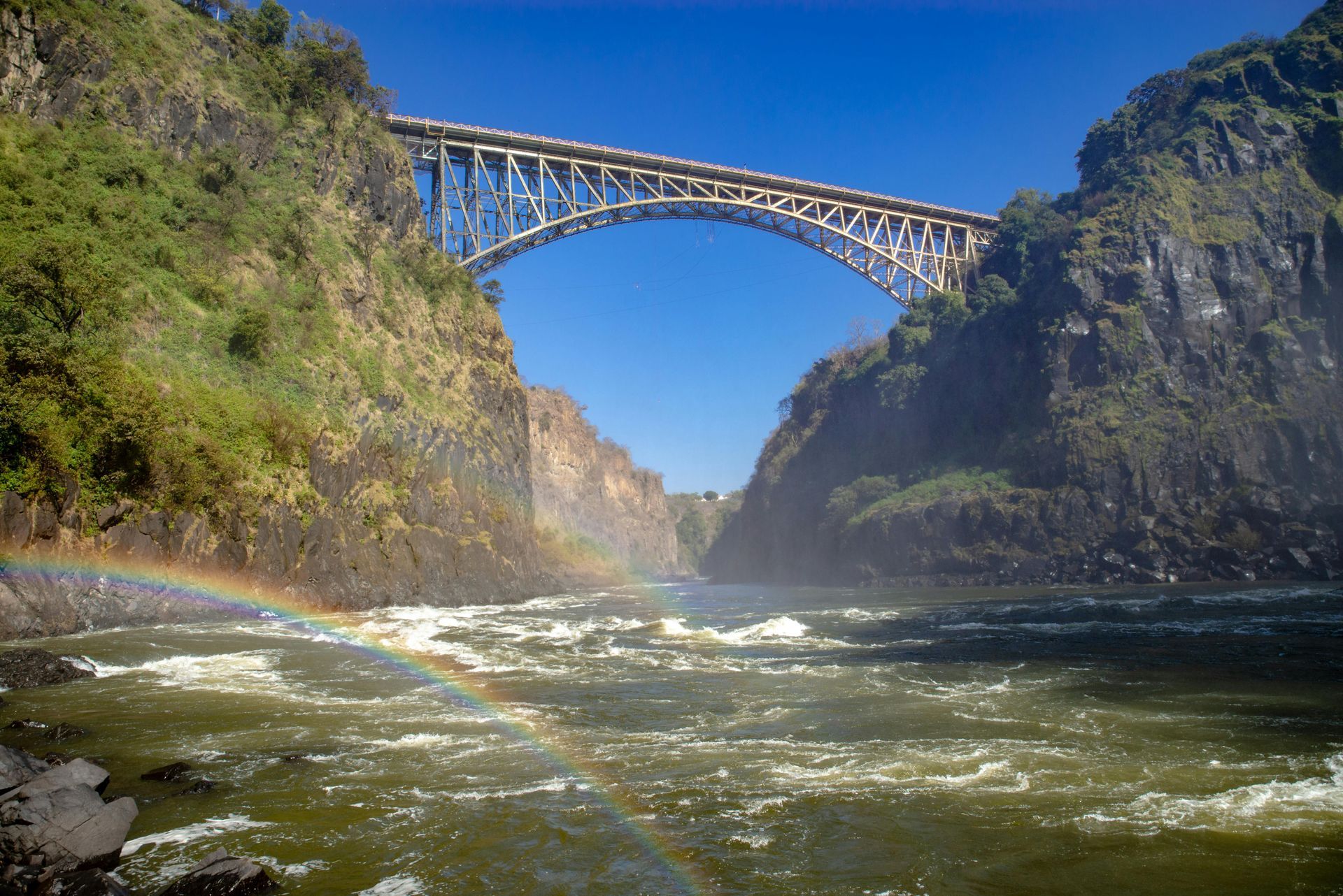 Bridge over a gorge with a rainbow, blue sky, and rushing water.