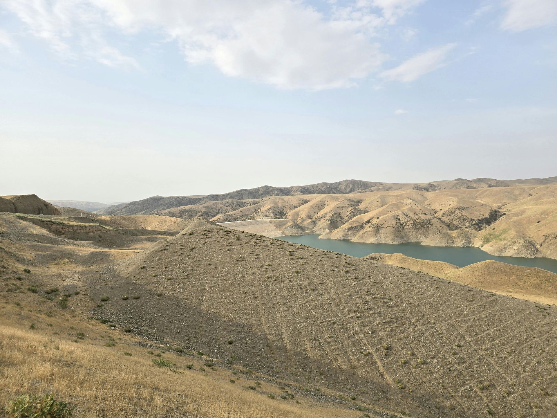 Hilly, arid landscape with a lake in the distance, under a partly cloudy sky.