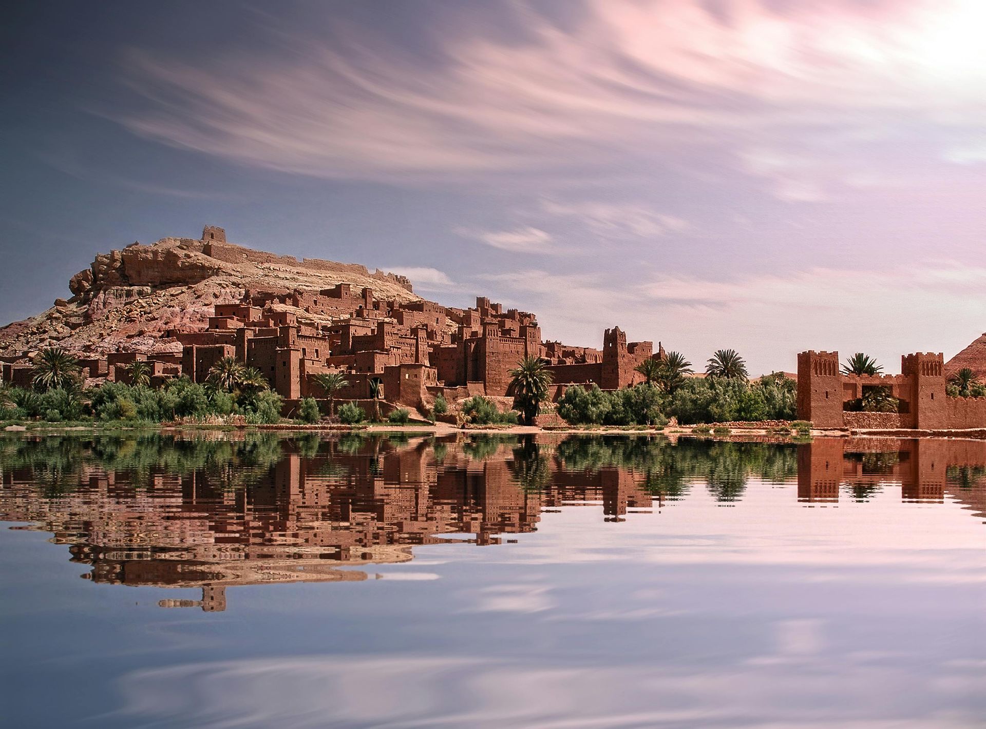 Ancient mud-brick buildings reflected in calm water under a pink-tinged sky.