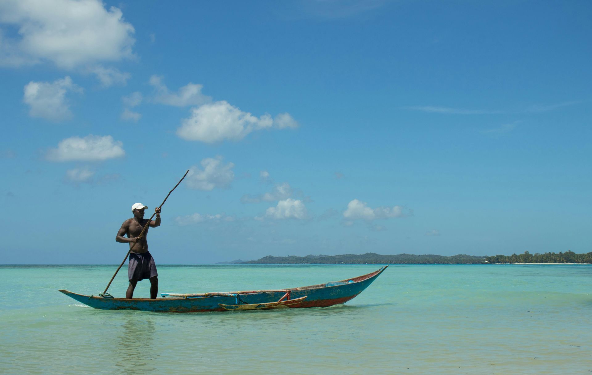 Man in a boat using a pole on turquoise water under a blue sky.