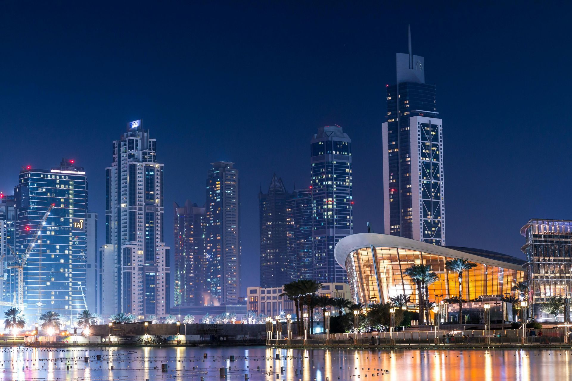 Nighttime skyline of Dubai, UAE, with illuminated skyscrapers reflecting in calm water under a dark blue sky.