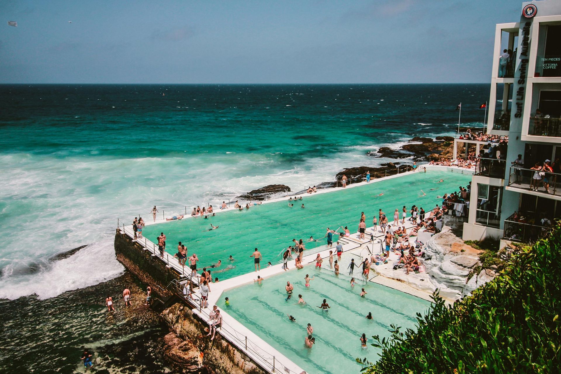 Pool with people next to ocean, white building on the right, blue-green water.