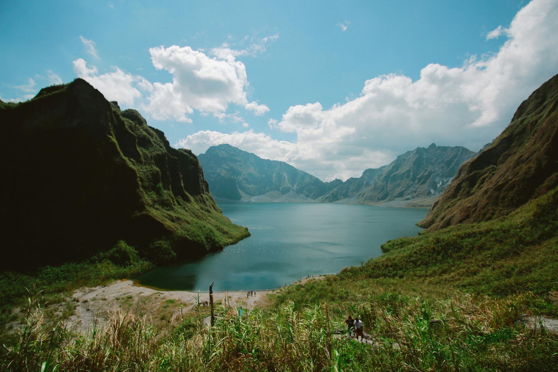 Lake surrounded by mountains under a blue sky with fluffy clouds; lush green vegetation in the foreground.
