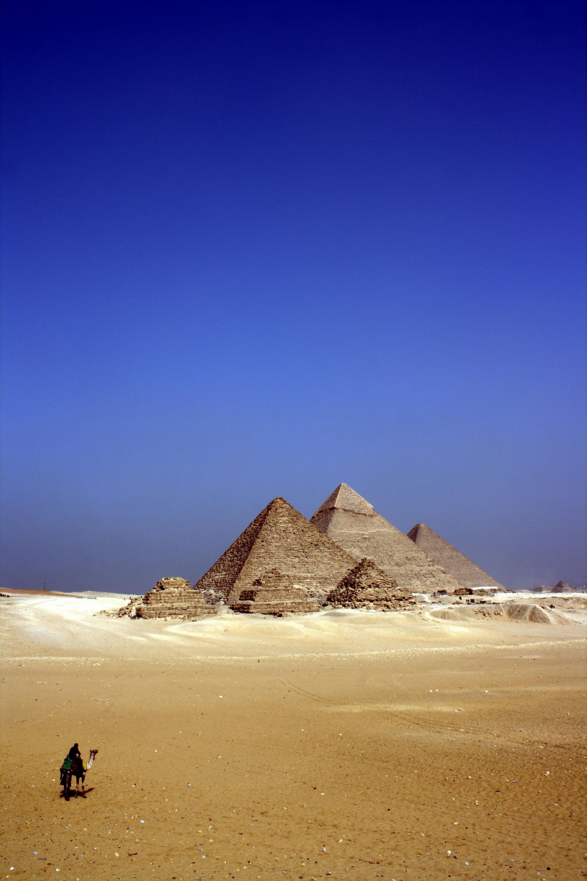 Pyramids of Giza in Egypt, with a lone figure on a camel in the foreground under a bright blue sky.