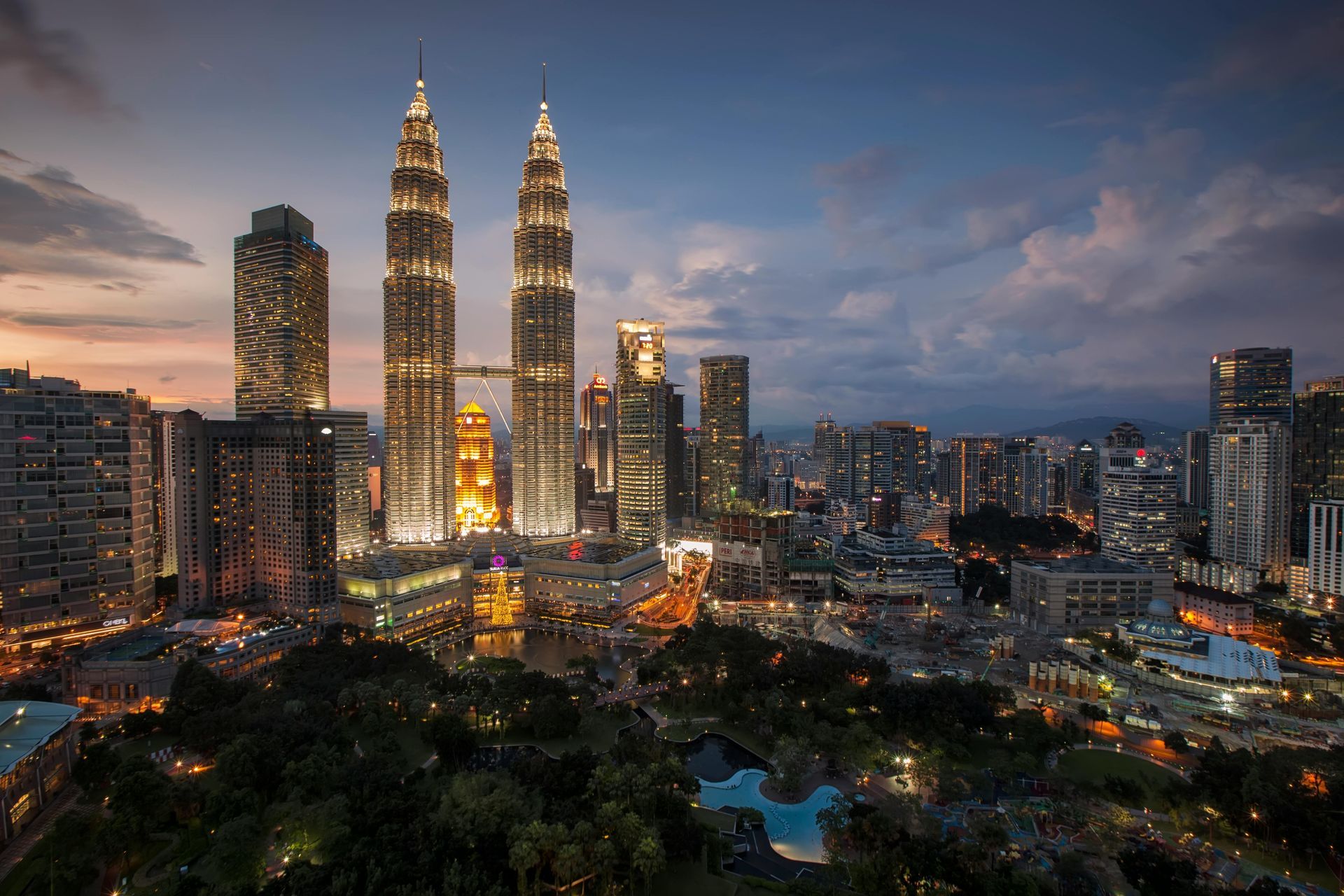 Kuala Lumpur skyline at dusk, featuring Petronas Towers. City lights illuminate buildings against a colorful sky.