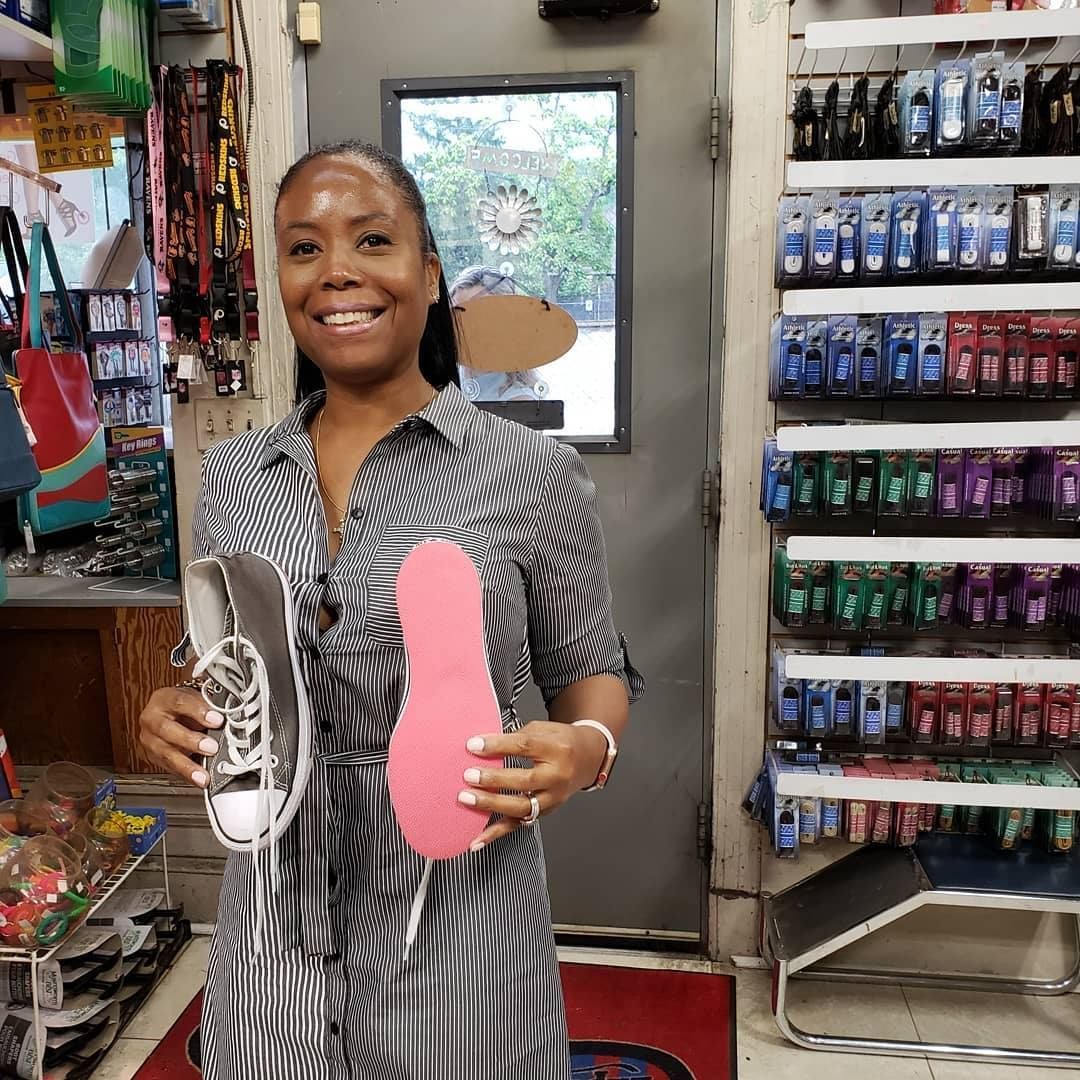 Woman in dress holds a shoe and pink insole in a shop; smiling, shelves of products.