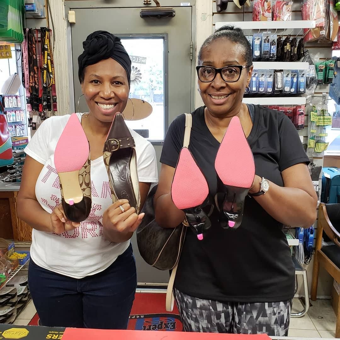 Two smiling women hold shoes with pink soles inside a shoe repair shop.