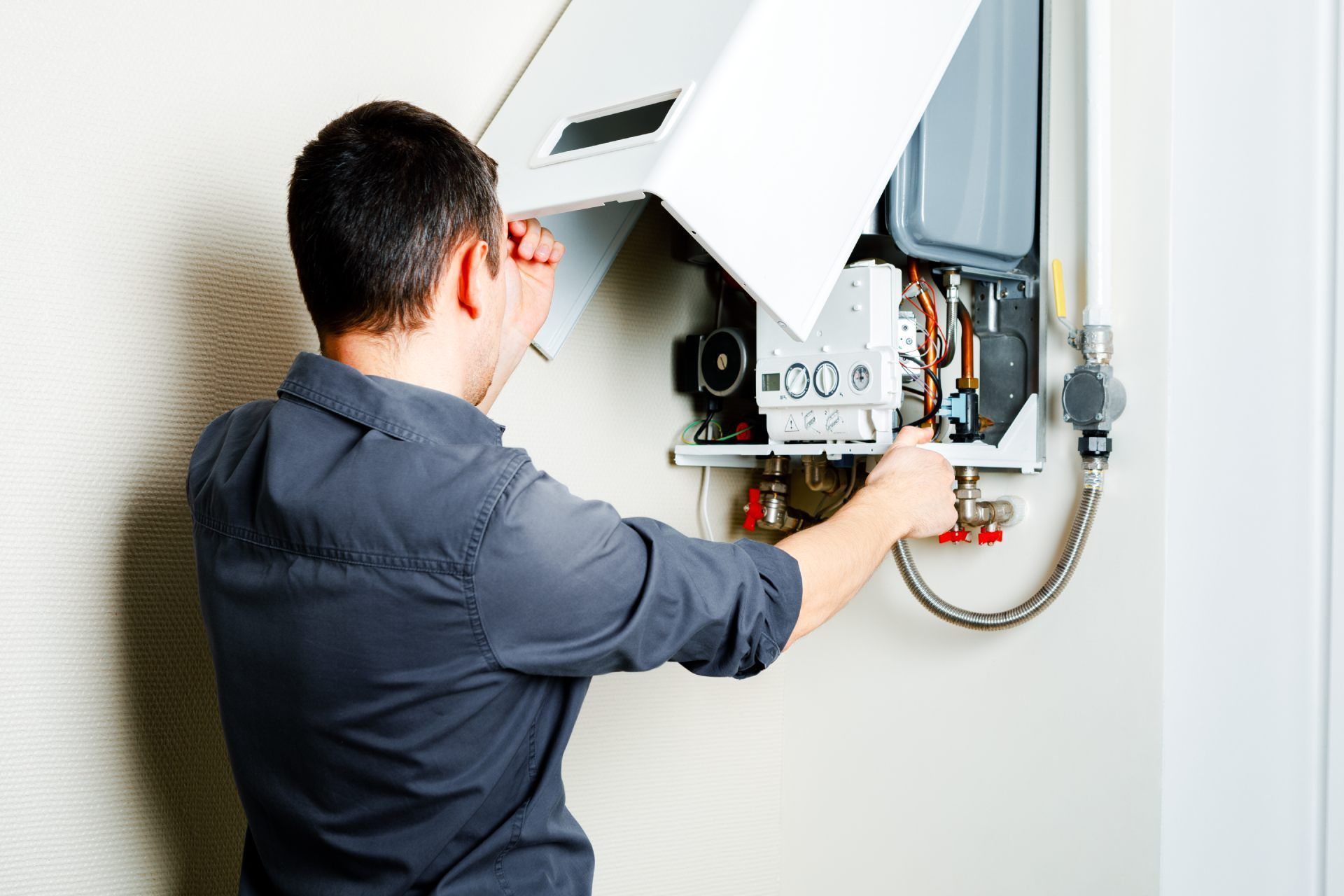 Man in gray shirt inspecting a white wall-mounted boiler, its access panel open.