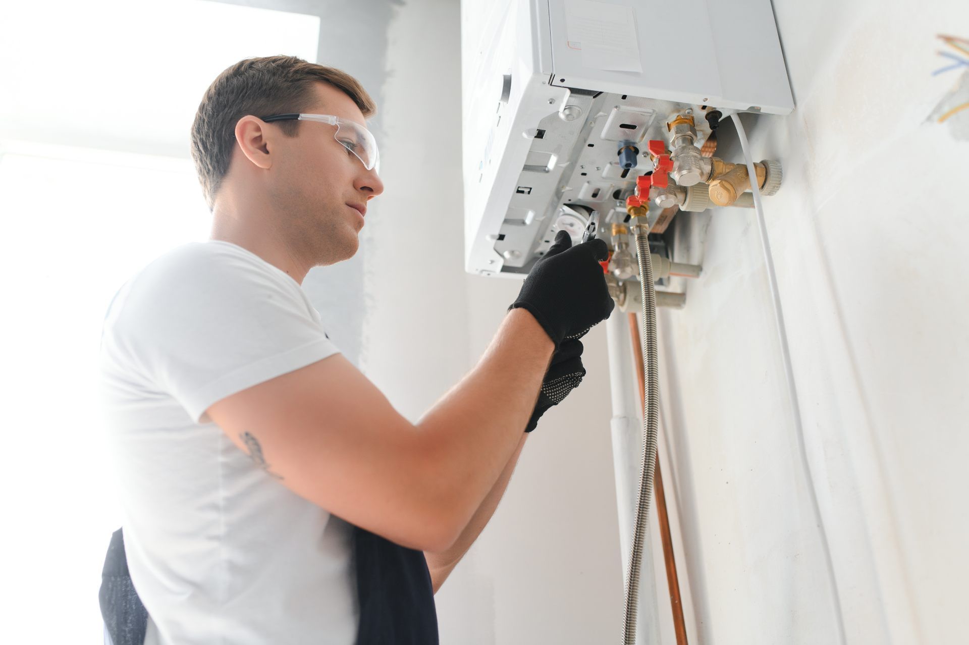 Person in safety glasses and gloves repairs a white appliance mounted on a wall.