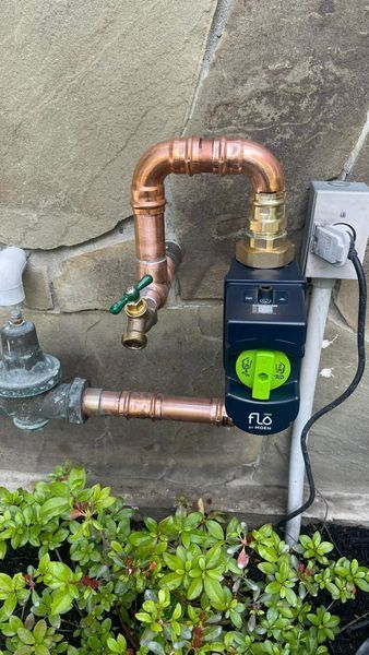 Copper water pipes with a green-accented water sensor attached to a stone wall, next to a bush.