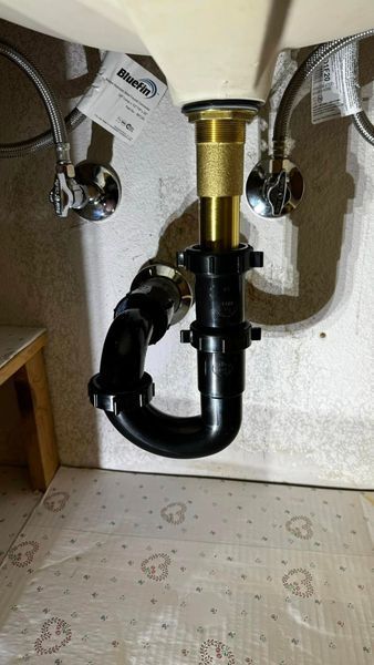 Under-sink plumbing: brass drainpipe, black p-trap, connected to water supply lines and a beige sink in a basement.