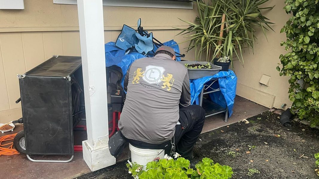 Person squatting next to a white bucket, working outdoors under a covered area, surrounded by equipment and tarps.