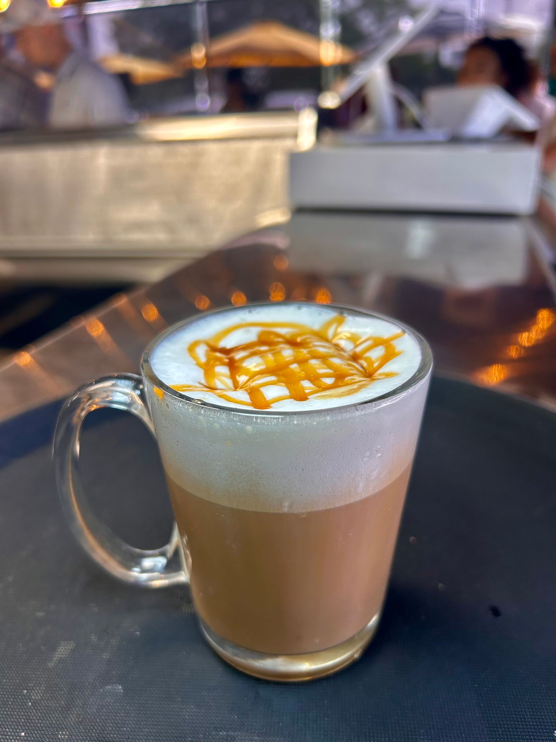 Caramel latte in a clear glass mug on a table, topped with foam and caramel drizzle.
