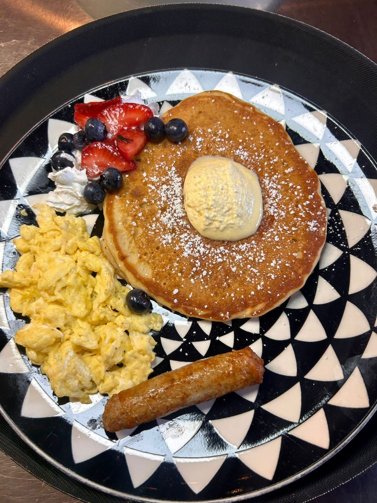 Pancakes with butter, blueberries, strawberries, whipped cream, scrambled eggs, and sausage on a patterned plate.