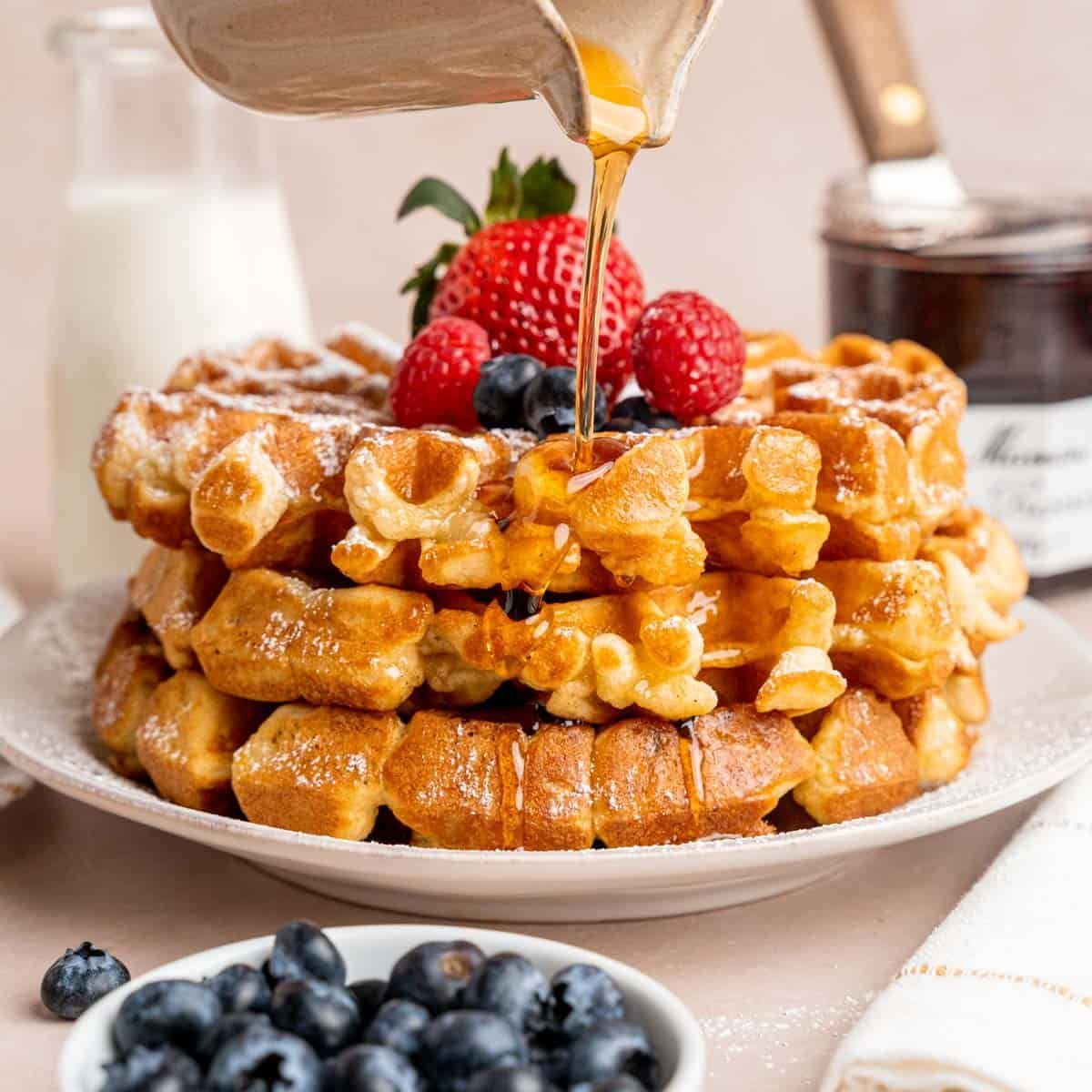 Syrup is being poured on a stack of waffles with strawberries and blueberries.