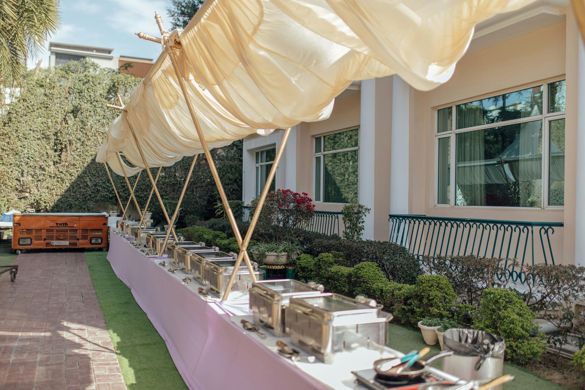 Outdoor buffet setup under a cream canopy beside a building, with serving dishes and plants along the walkway