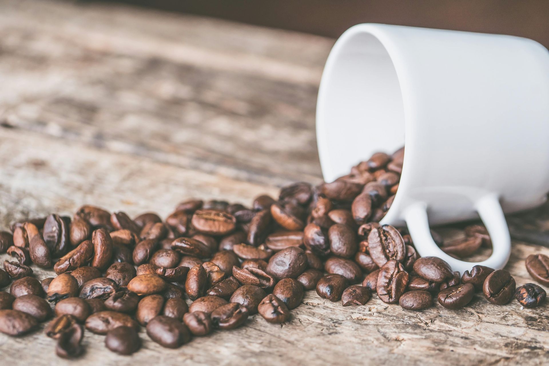White mug spilling roasted coffee beans onto a rustic wooden surface.