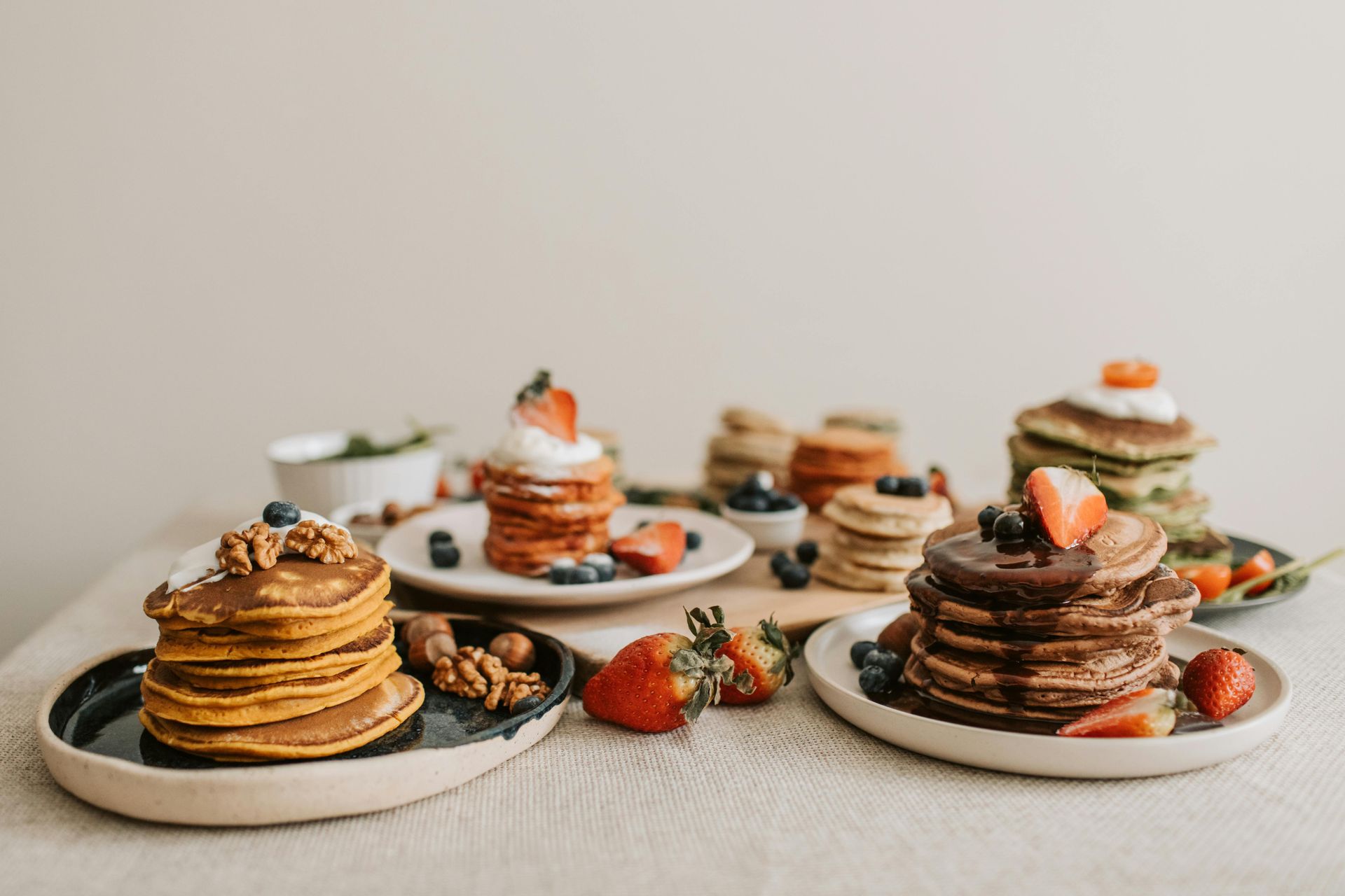 Stacks of colorful pancakes with toppings like fruit, berries, and nuts on plates, set on a table.