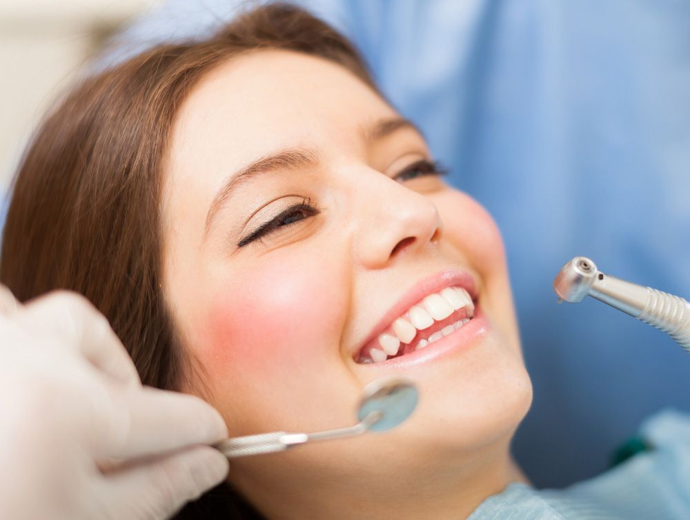 Woman smiling at dentist, in dentist's chair. Dentist's tools are visible, pink cheeks.