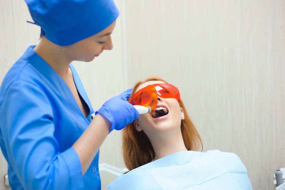 Dentist using a light on a patient’s teeth, patient wears orange goggles, inside dental office.