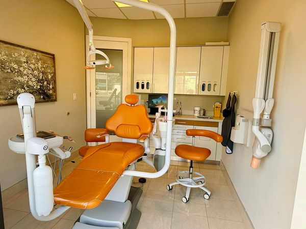 Orange dental chair in a dental office with cabinets, a dental light, and a wall-mounted X-ray machine.