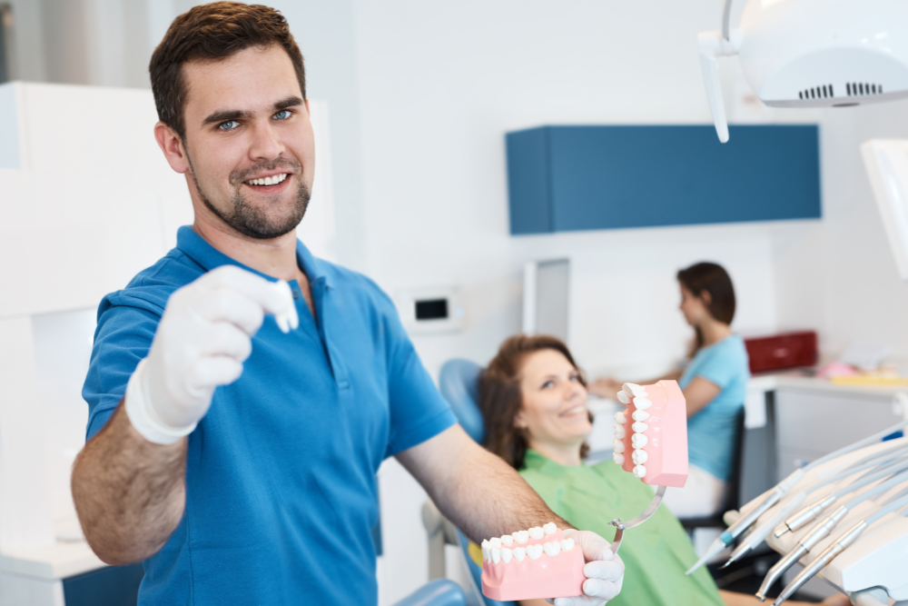 Dentist holding a tooth and teeth model, smiling in dental office with patient.