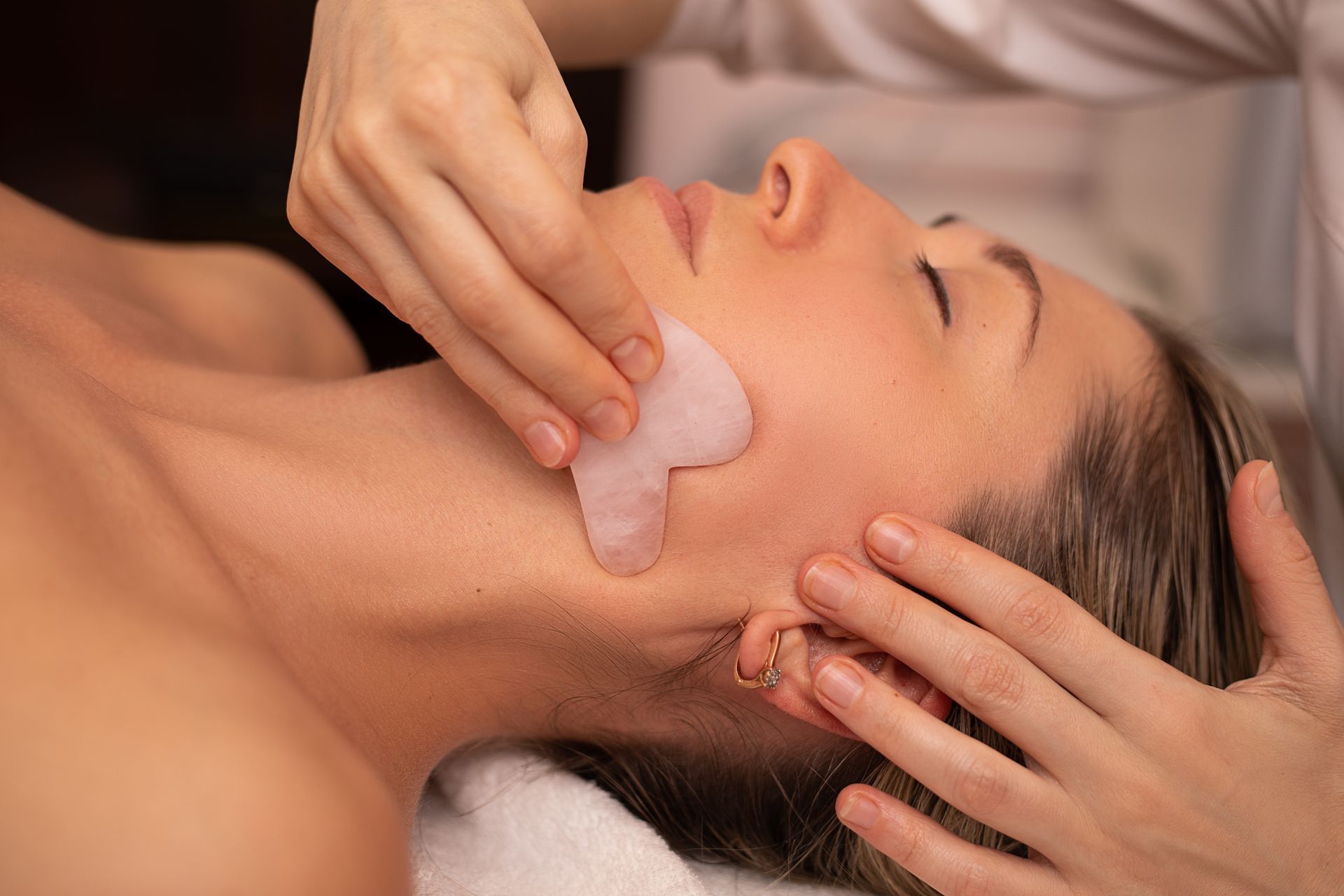 Gesichtsmassage mit beiden Händen, bei der Creme auf das Gesicht einer liegenden Person aufgetragen wird