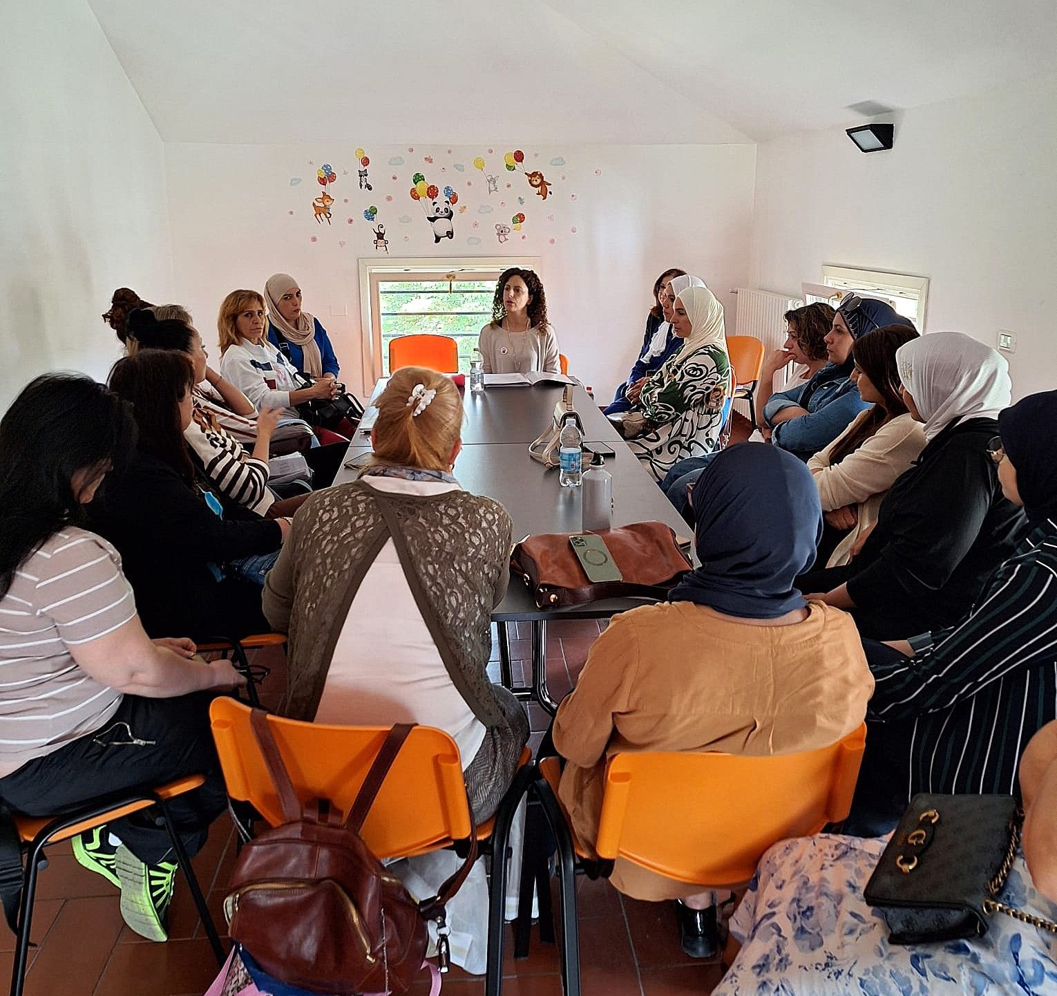 Un gruppo di persone sedute attorno a un tavolo in una stanza ascolta una donna che parla.