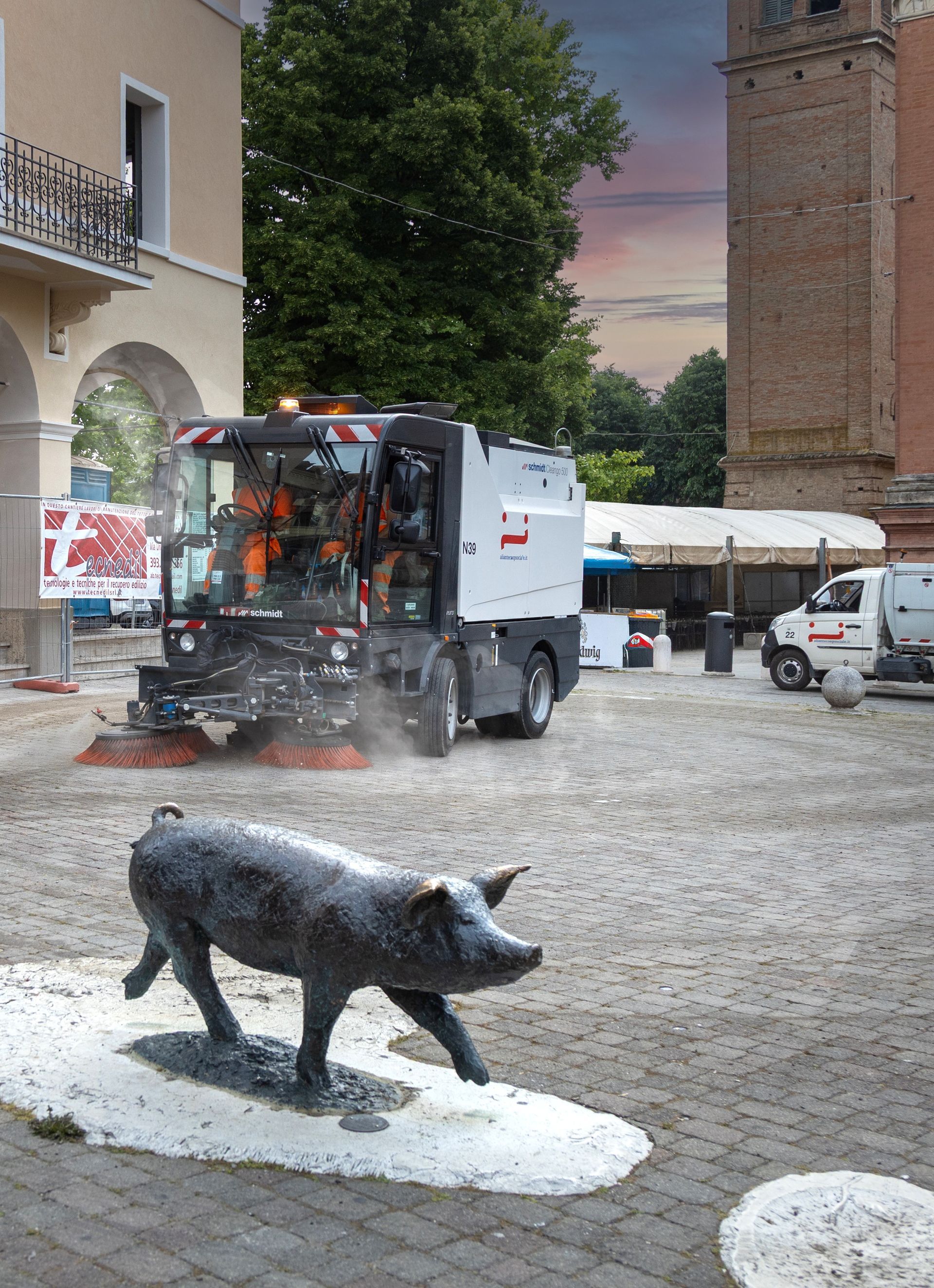 Spazzino che pulisce una piazza pubblica, con la statua di un maiale in primo piano.