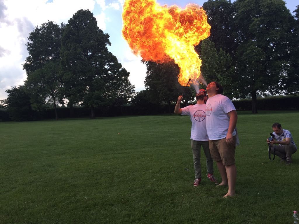 Due persone soffiano sul fuoco in un campo erboso, una delle quali esala una grande fiamma. Il fotografo è inginocchiato lì vicino.